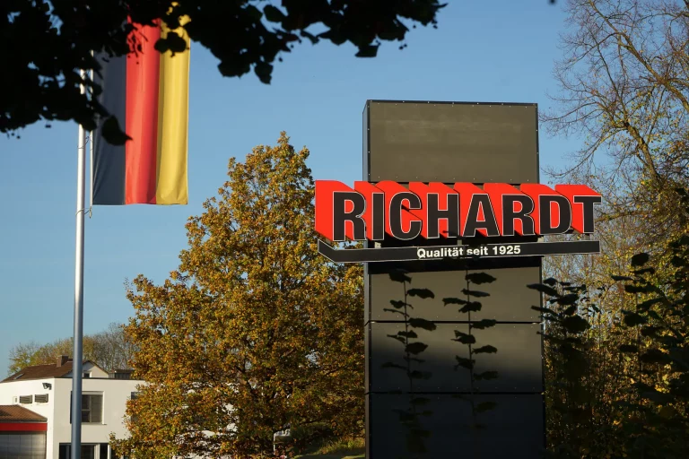 Außenansicht des Firmenschilds „Richardt – Qualität seit 1925“ neben deutscher Flagge und Herbstbäumen.
